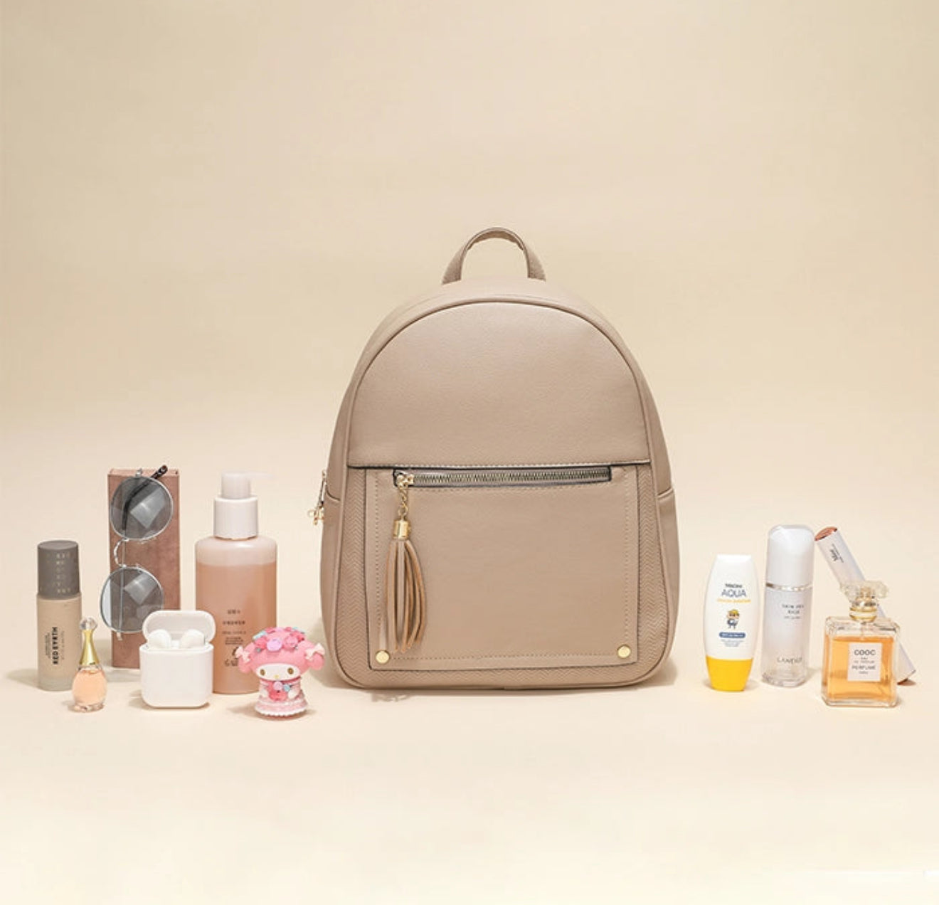 A beige faux-leather backpack with a front zip pocket and a tassel zipper pull, styled in a flat-lay arrangement with various beauty and perfume products on a warm beige background.