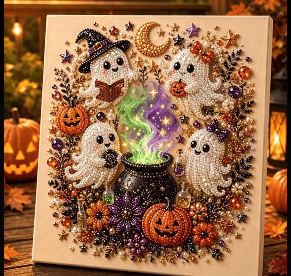 A square Halloween-themed beaded artwork or craft panel featuring four smiling ghosts, a black cauldron with glowing green and purple smoke, orange pumpkins, stars, moons, and floral accents on a cream background, displayed on a wooden surface with warm autumn decor blurred in the background.