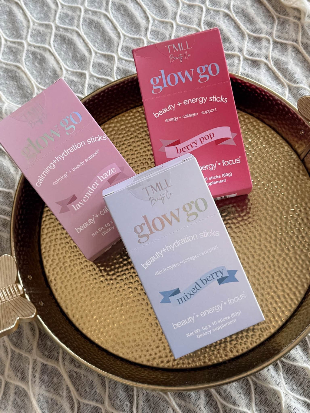 Three boxes of TMLL Bright glow go beauty + energy sticks arranged on a round gold tray over a textured white fabric background. The boxes are pink, coral pink, and light lavender, with the text 'TMLL Bright glow go' and flavor names 'berry bud', 'lavender hint', and 'mixed berry' visible.