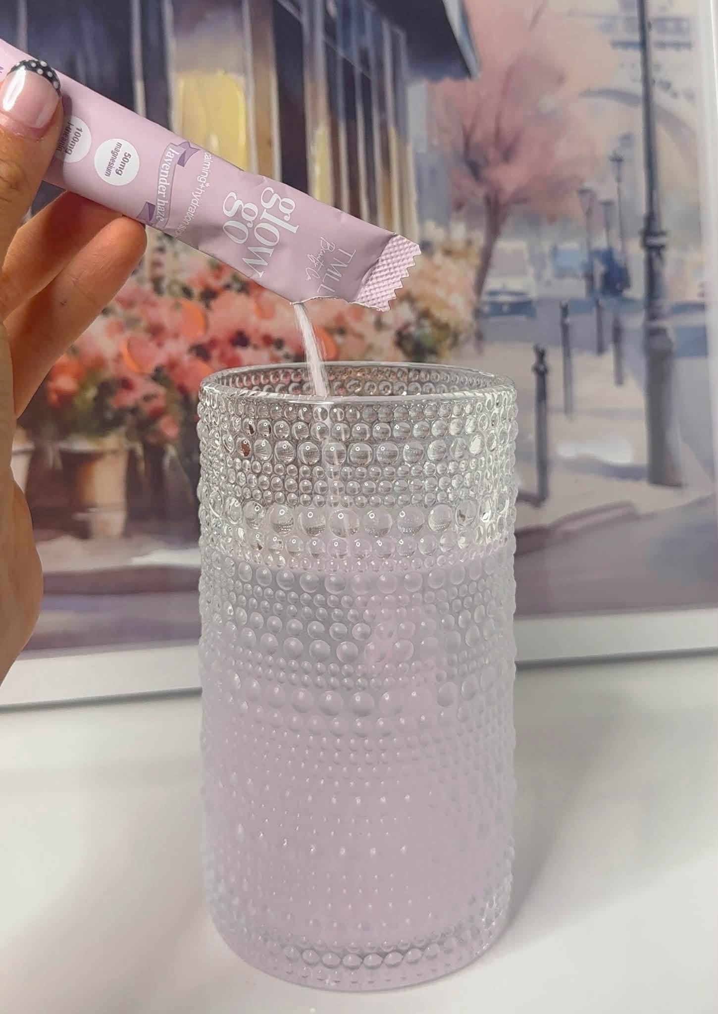 A hand holding a pink stick packet above a textured clear glass cup, with a pale pink liquid being poured into the cup. The background shows a blurred framed picture of a street scene.