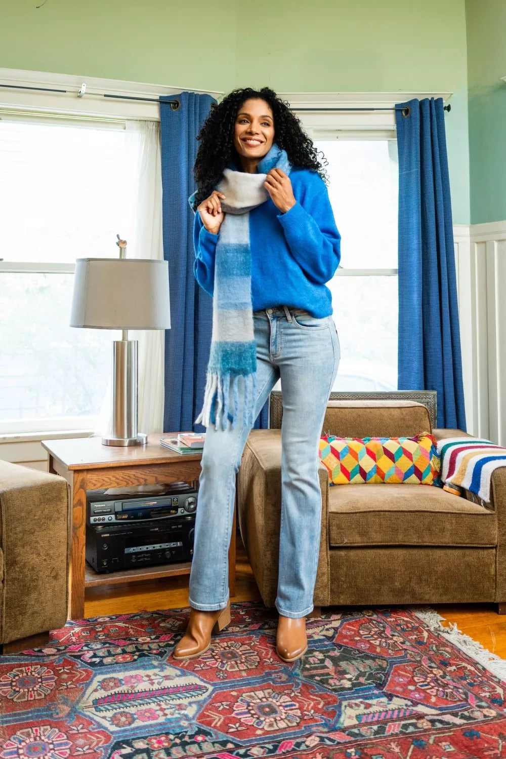 Woman in a blue sweater and scarf standing in a living room with a colorful rug and furniture.