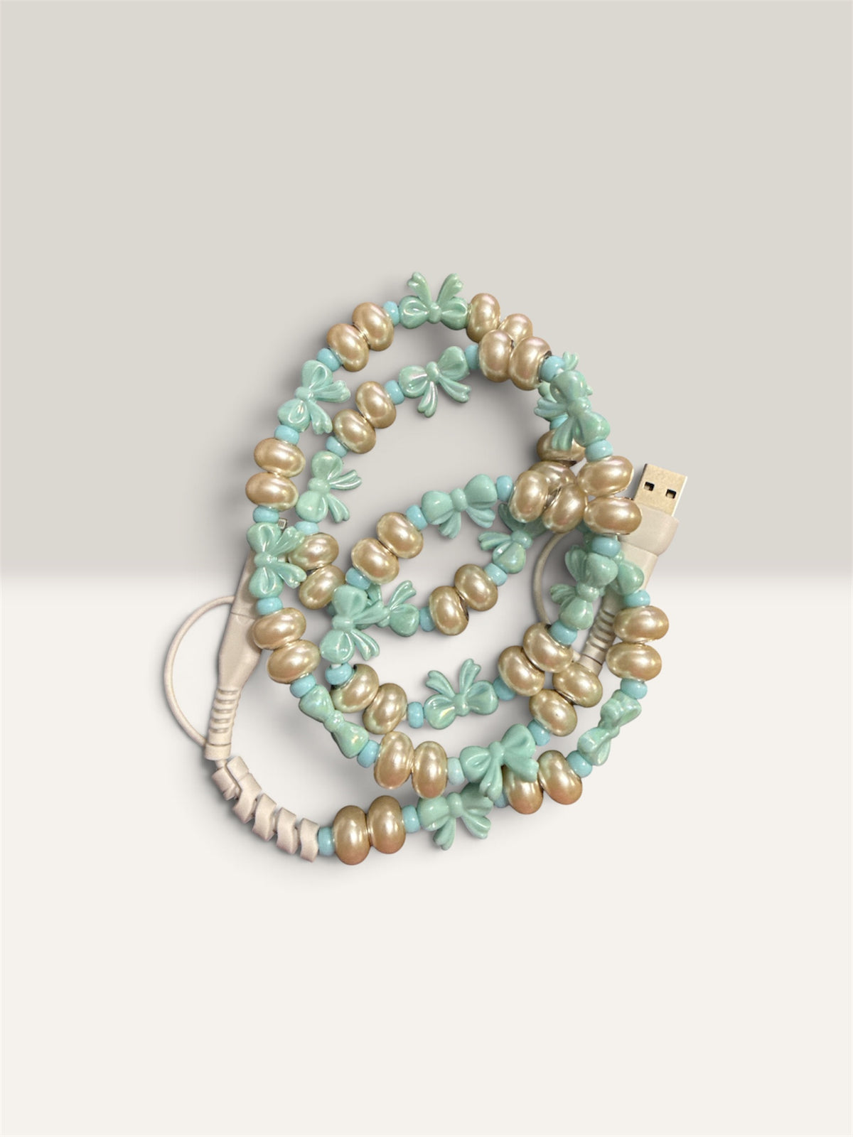 A coiled USB charging cable decorated with light green faux flowers and beige faux pearls, shown on a neutral light gray background.