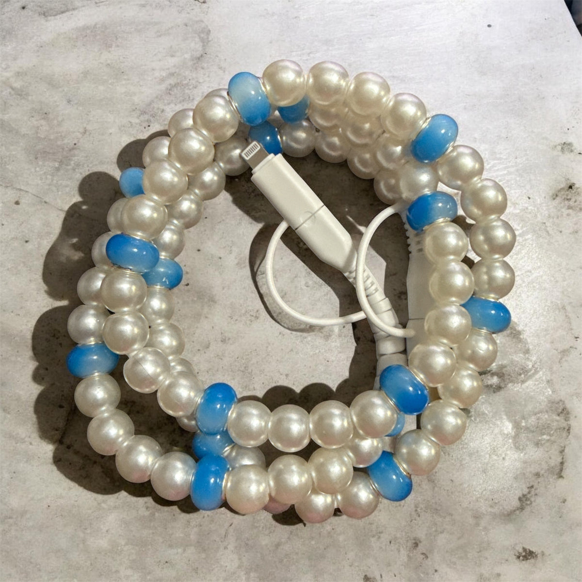 A coiled beaded charger made of mostly white faux pearls with scattered translucent light blue beads, shown on a light stone surface with a white charging cable placed over it.