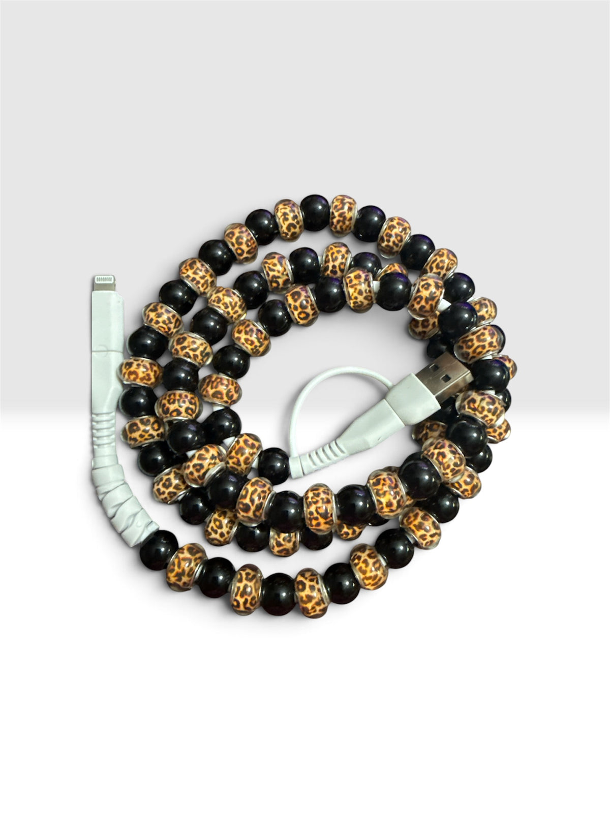 A coiled beaded phone cable featuring alternating black beads and leopard-print accent beads in brown and beige, with white lightning and USB connectors, shown on a light gray background.