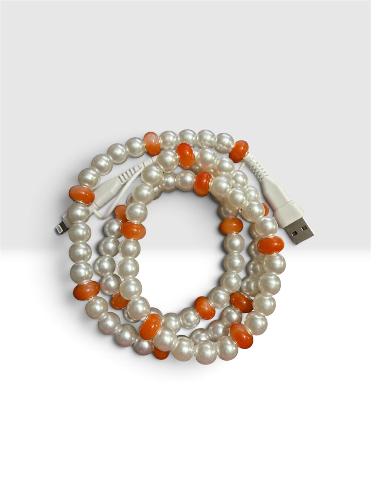 A coiled multi-strand pearl necklace with alternating white pearls and smooth orange beads, attached to a white USB charging cable, shown on a light gray background.