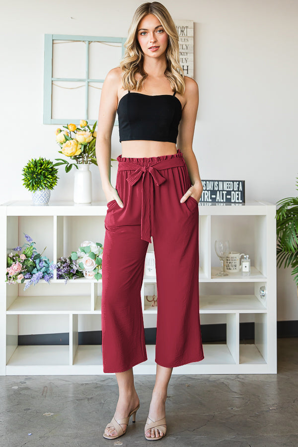 Woman wearing a black top and red high-waisted pants standing in a room with white shelves and decor.