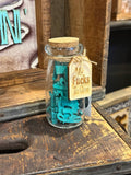 A small clear glass jar with a cork stopper sits on a rustic wooden surface. Inside the jar are bright teal letter tiles, and a tan paper tag tied with twine hangs from the rim.