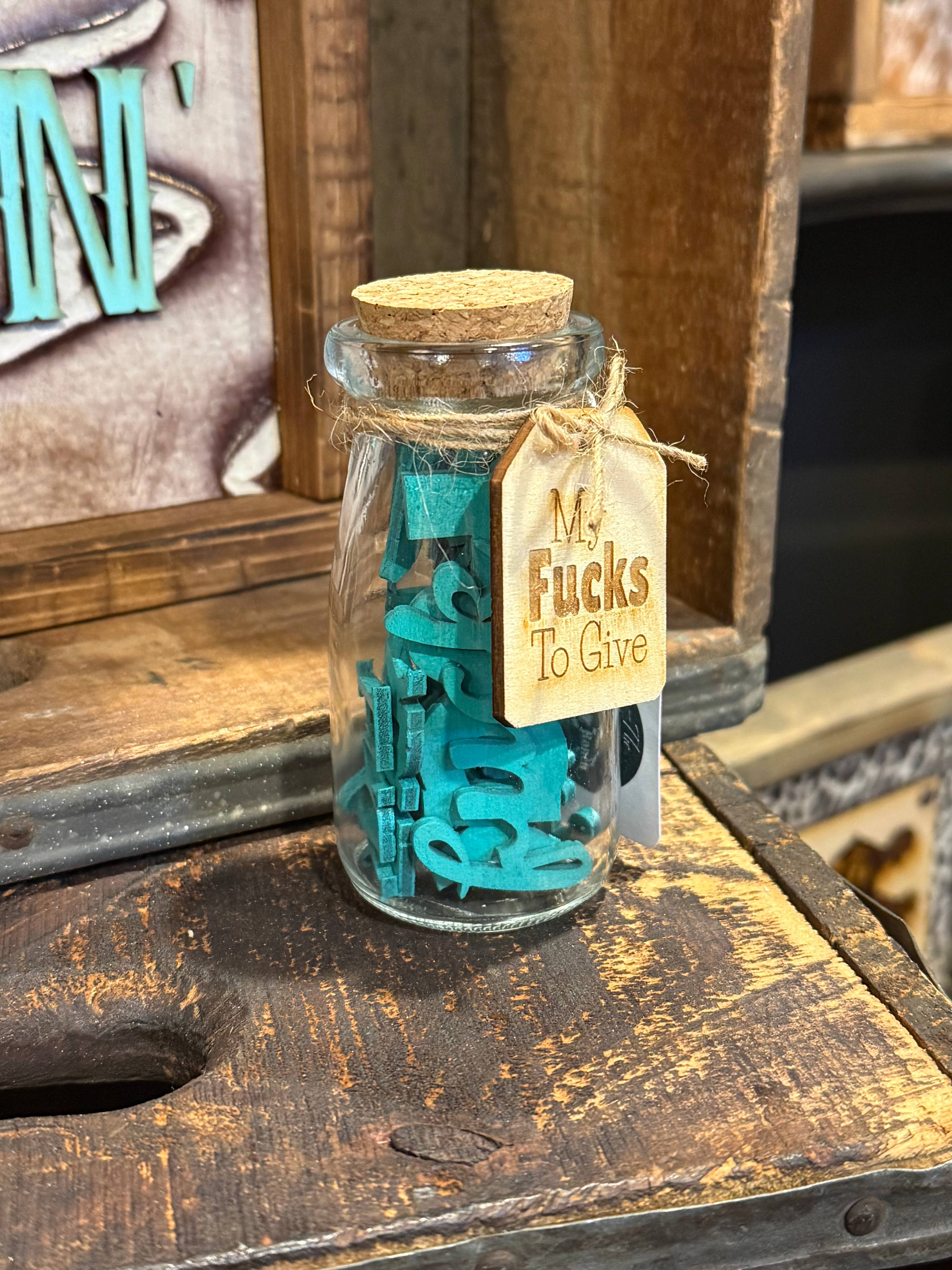 A small clear glass jar with a cork stopper sits on a rustic wooden surface. Inside the jar are bright teal letter tiles, and a tan paper tag tied with twine hangs from the rim.
