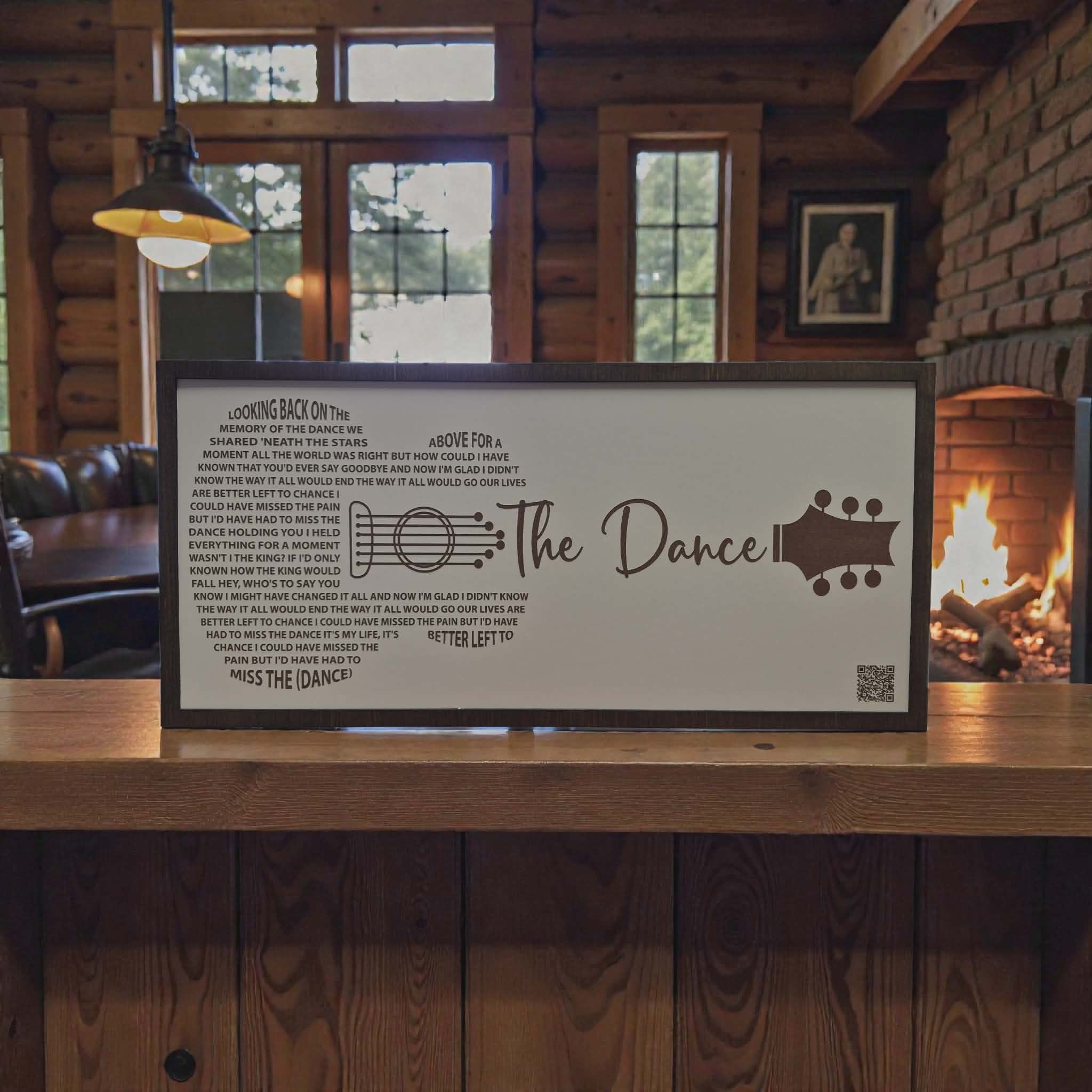 A framed wall sign featuring the lyrics and guitar graphic from the song 'Looking On The Bright Side Of Life' by The Dance, displayed on a wooden mantel in a cozy log cabin living room with a fireplace in the background.