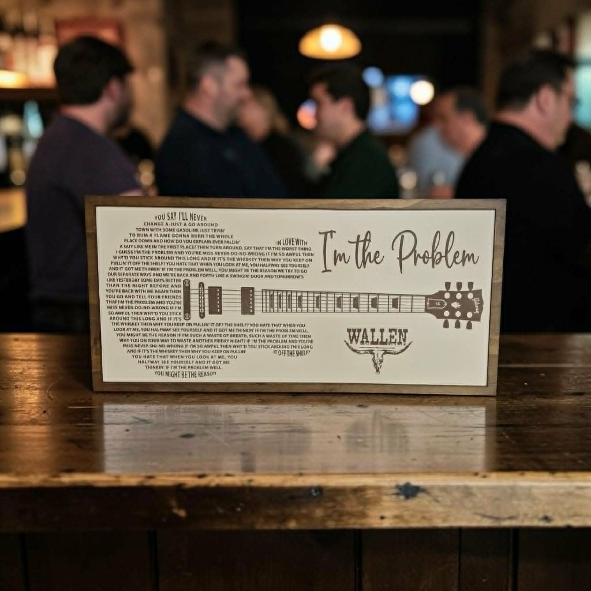 A framed wall sign featuring the song 'I'm the Problem' by Wallen, displayed on a rustic wooden bar counter with a blurred bar scene in the background.
