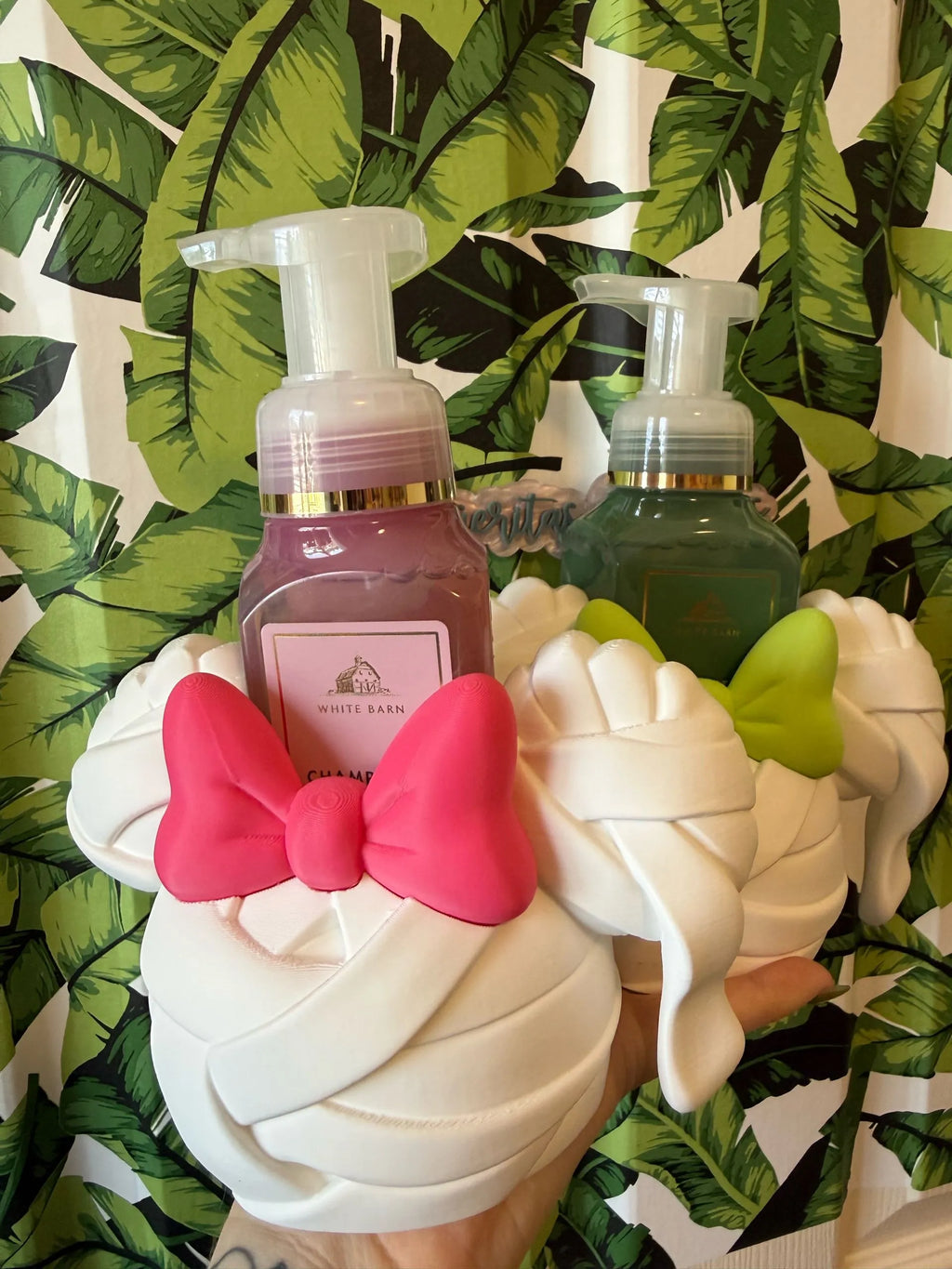 A hand holding two decorative pump bottles shaped like Minnie Mouse ears, with one bottle filled with pink liquid and the other with green liquid, both decorated with white faux-fur details and bright pink and green bows. A pink 'White Bean' label is visible on the front bottle.