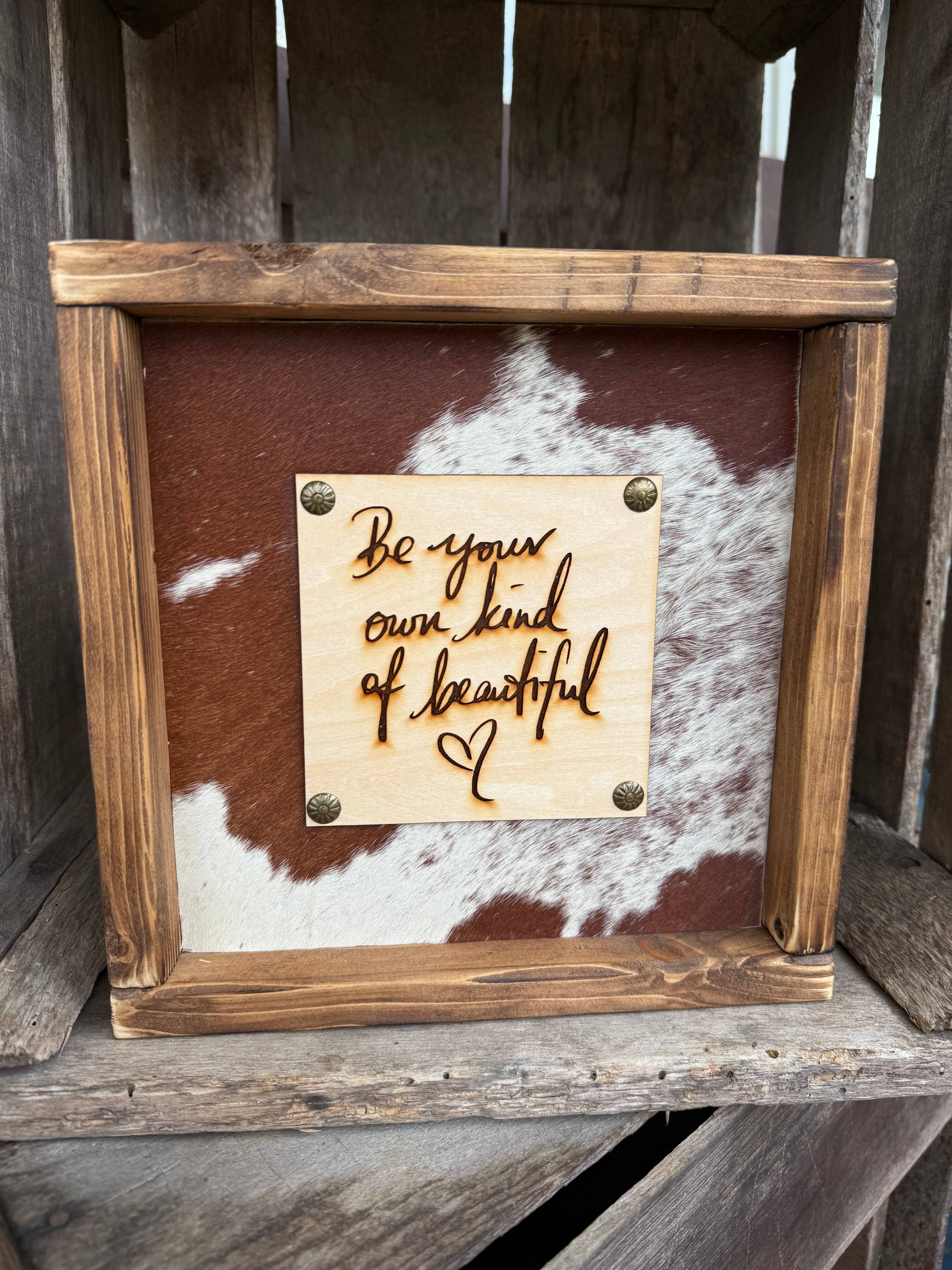 A rustic wooden framed sign featuring a cowhide background and a central wooden plaque with the engraved phrase 'Be your own kind of beautiful' and a small heart, displayed on a weathered wooden surface.
