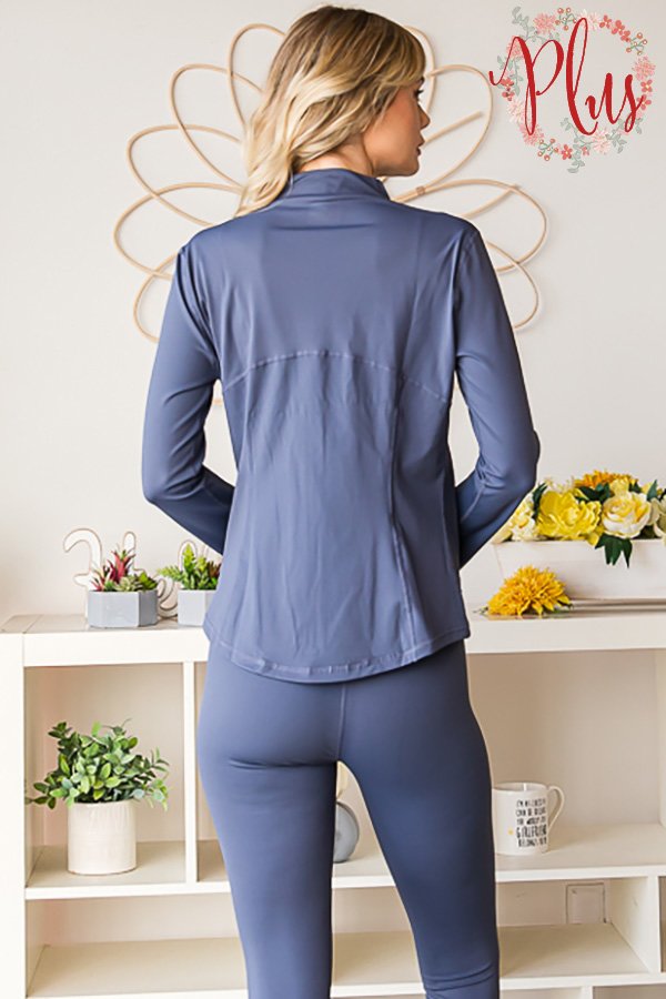 A woman shown from the back wearing a matching slate blue athletic set consisting of a long-sleeve zip-up jacket and high-waisted leggings, standing in front of a white shelf with decorative plants and flowers.